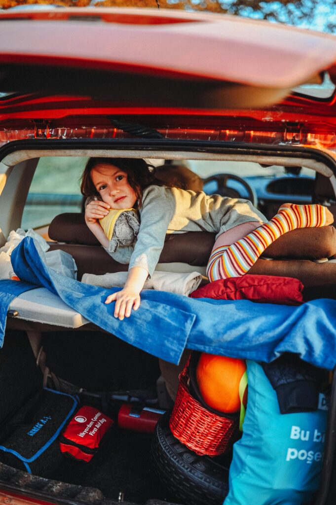 A young girl lying in the trunk of a car, surrounded by travel bags and gear, ready for an adventure.